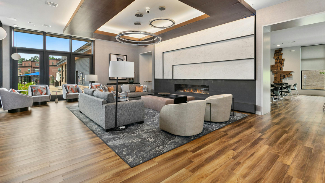 A modern, spacious residential lounge with wood floors, gray furniture, a large fireplace, and expansive windows. The interior design features contemporary lighting fixtures.
