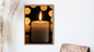 Close-up photographic wall art of a thick, white pillar candle with a bright, steady flame. The background is dark, featuring numerous large, warm, out-of-focus golden circles (bokeh) surrounding the candle. The art is framed in light wood and mounted on a white wall. Wall art frame in a living room by wall and more