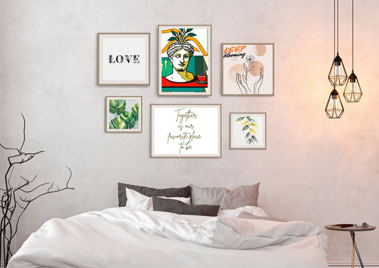 The gallery wall features six framed artworks in natural wood frames, including: An abstract portrait of a classical bust in vibrant yellow, red, and turquoise. Minimalist black typography stating "LOVE." A hand-drawn sketch of hands holding a flower with "KEEP blooming." A square print of lush green tropical leaves. A small print of stylized yellow and green leaves. Flowing gold calligraphy stating "Together is our favourite place to be.".Wall gallery art by wall and more